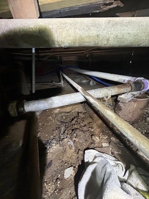 Pipes and wooden beams in a dimly lit crawl space. White, gray, and blue pipes sit in dirt.