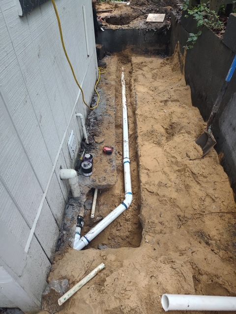 Trench with white plumbing pipes along a wall. Construction scene, dirt, tools, and a dark shaded area in the background.