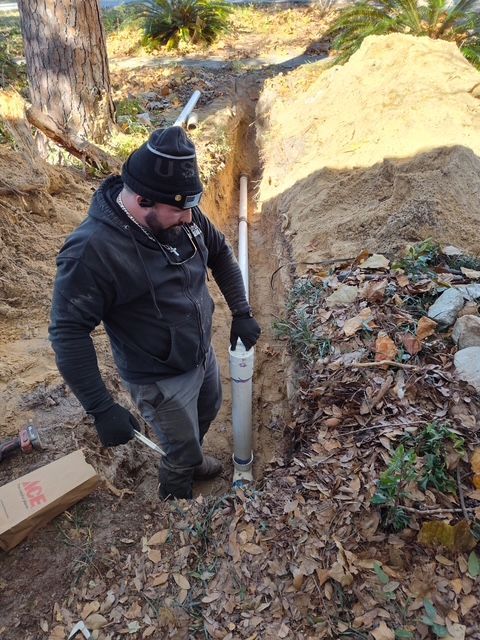 Man installing a PVC pipe in a trench outdoors, wearing a hooded jacket and gloves.