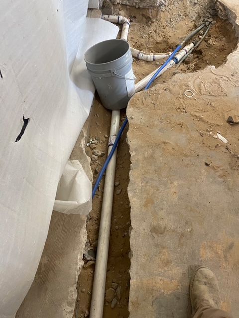 Trench with plumbing pipes, bucket, blue wire, and white fabric on a concrete floor.