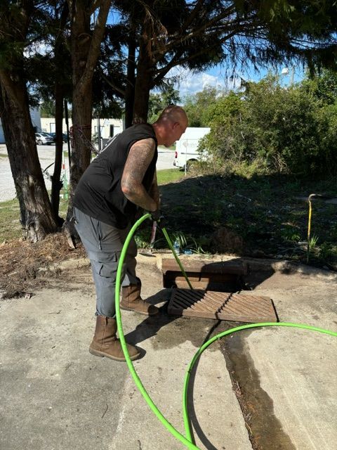 Man holding a green hose, washing a dark, square drain outdoors.