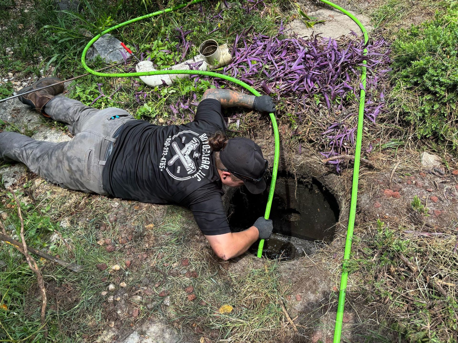 Man in black shirt, cap, and gloves working near open hole, holding green hose. Outdoors, near purple flowers.