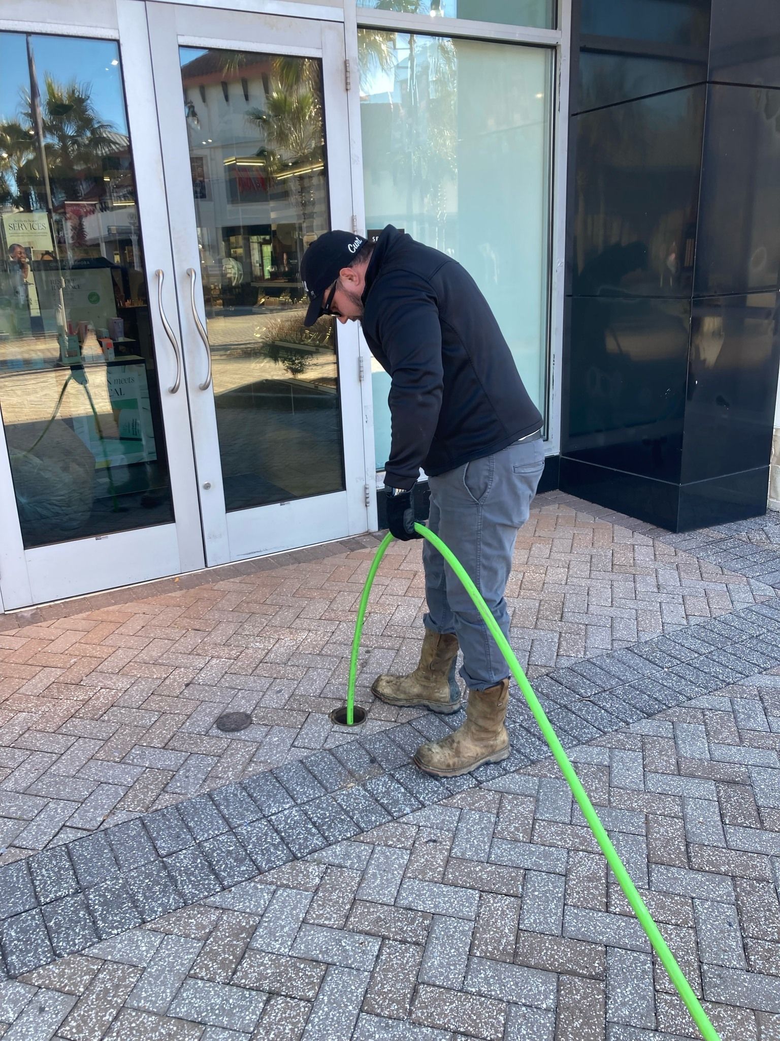 A person in work clothes uses a green hose to clean a drain on a brick sidewalk next to a building.