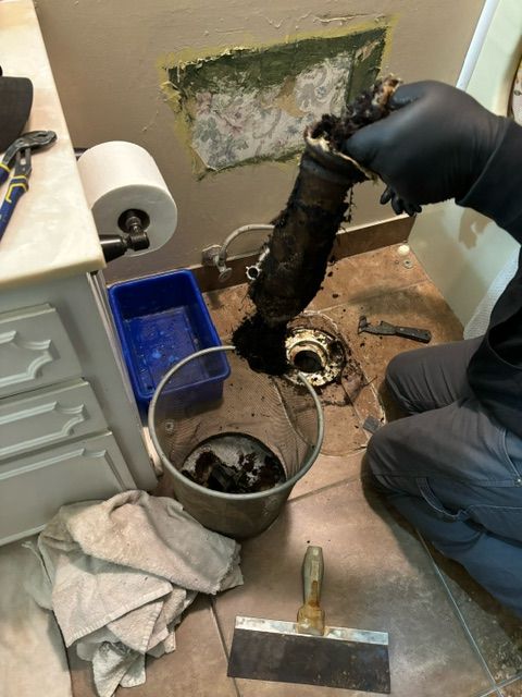 A person holding a clogged, dirty pipe in a bathroom; a bucket holds debris.