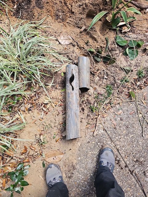 Broken gray pipe pieces on sandy ground. Feet in gray shoes are visible.