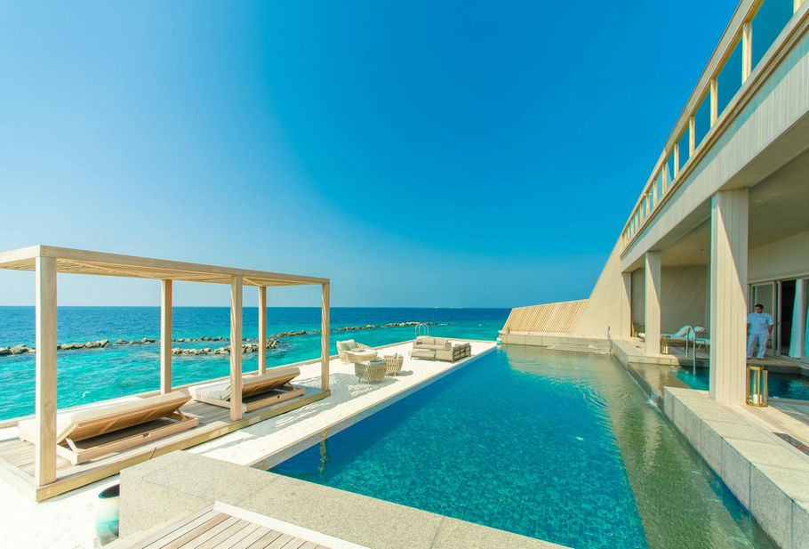 An infinity pool at a luxury resort overlooks a bright blue ocean under a clear sky, featuring wooden lounge furniture.