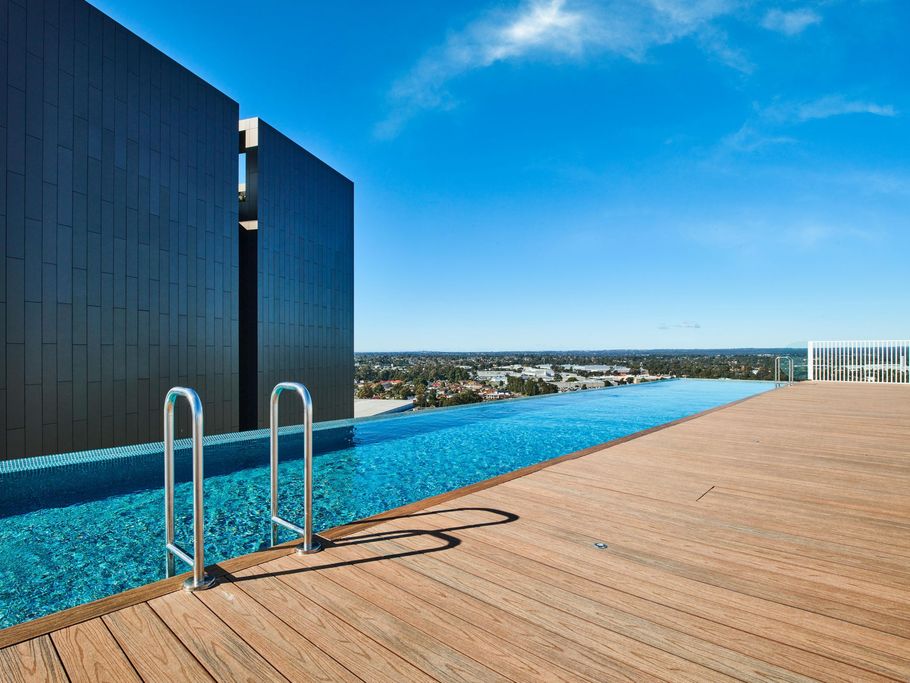 Infinity pool on a rooftop terrace with a wooden deck and a view of the city skyline under a clear blue sky.
