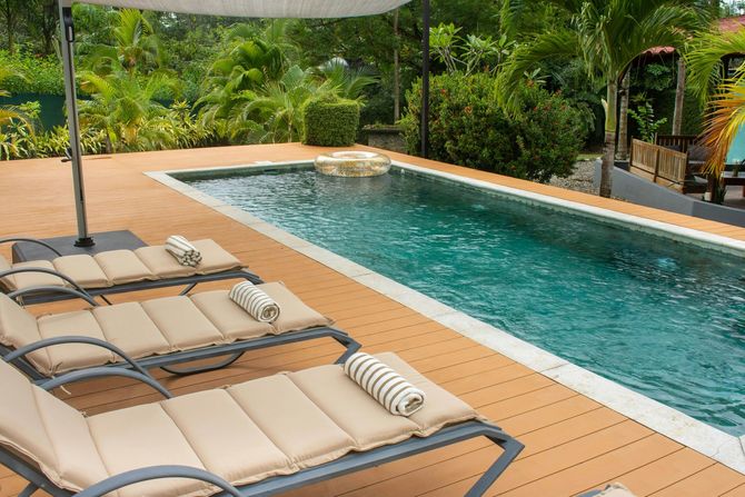 Three lounge chairs with striped pillows on a wooden pool deck next to a rectangular swimming pool surrounded by greenery.