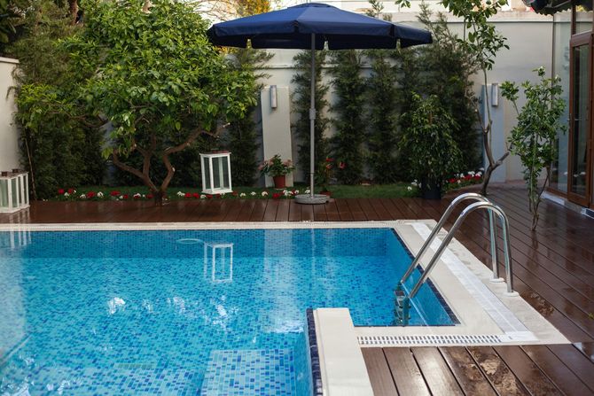 A rectangular blue tiled swimming pool with metal steps, set on a wooden deck next to a blue umbrella and lush green trees.