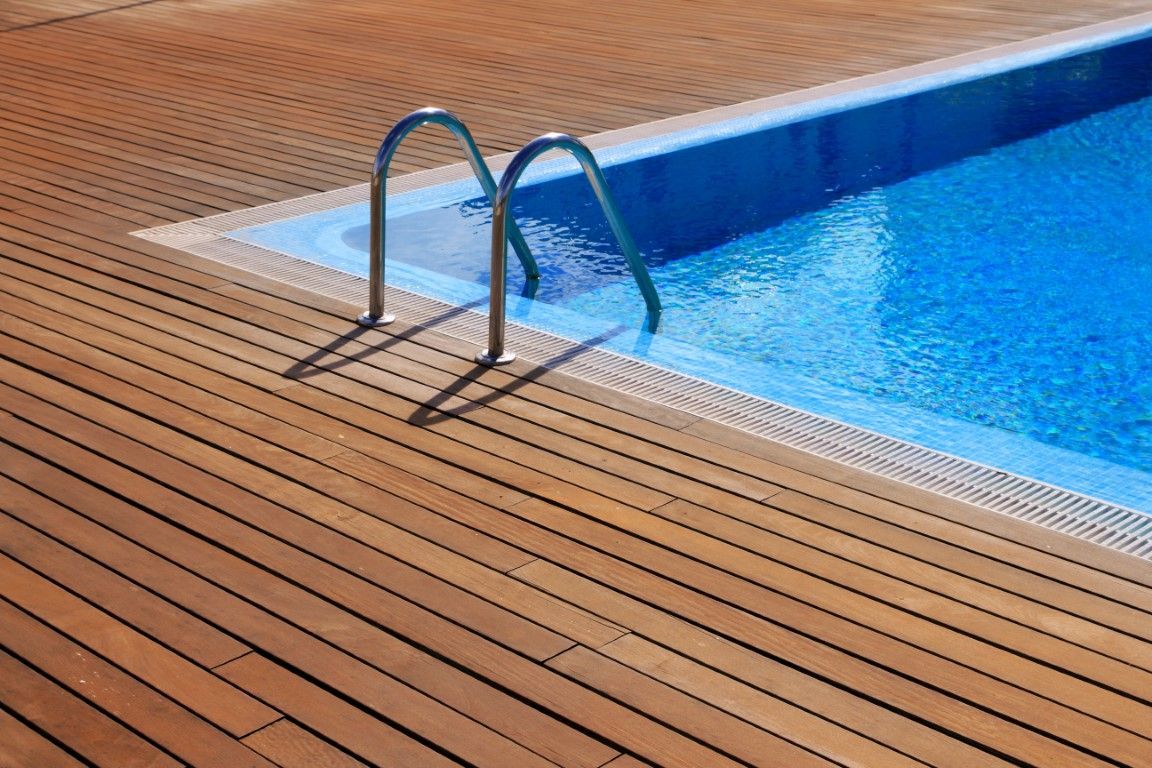 A top-down view of a clear blue swimming pool corner bordered by weathered, gray wooden decking and green grass.