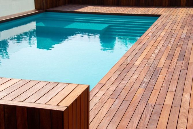 A sunny rectangular swimming pool with light blue water and stone tiling, surrounded by grass and trees.