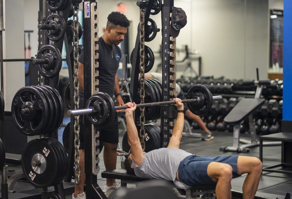Person bench pressing in a gym while a trainer spots nearby
