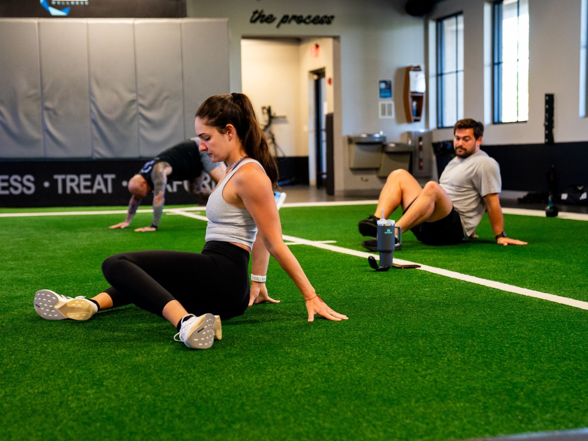 People stretching on green turf in FTX Performance in Boca Raton, FL, with exercise equipment and a trainer in the background
