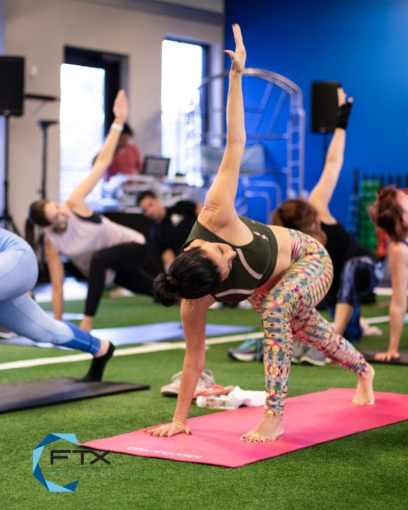 People doing yoga poses in FTX Performance in Boca Raton, FL on mats, with a woman balancing on a pink mat in the foreground
