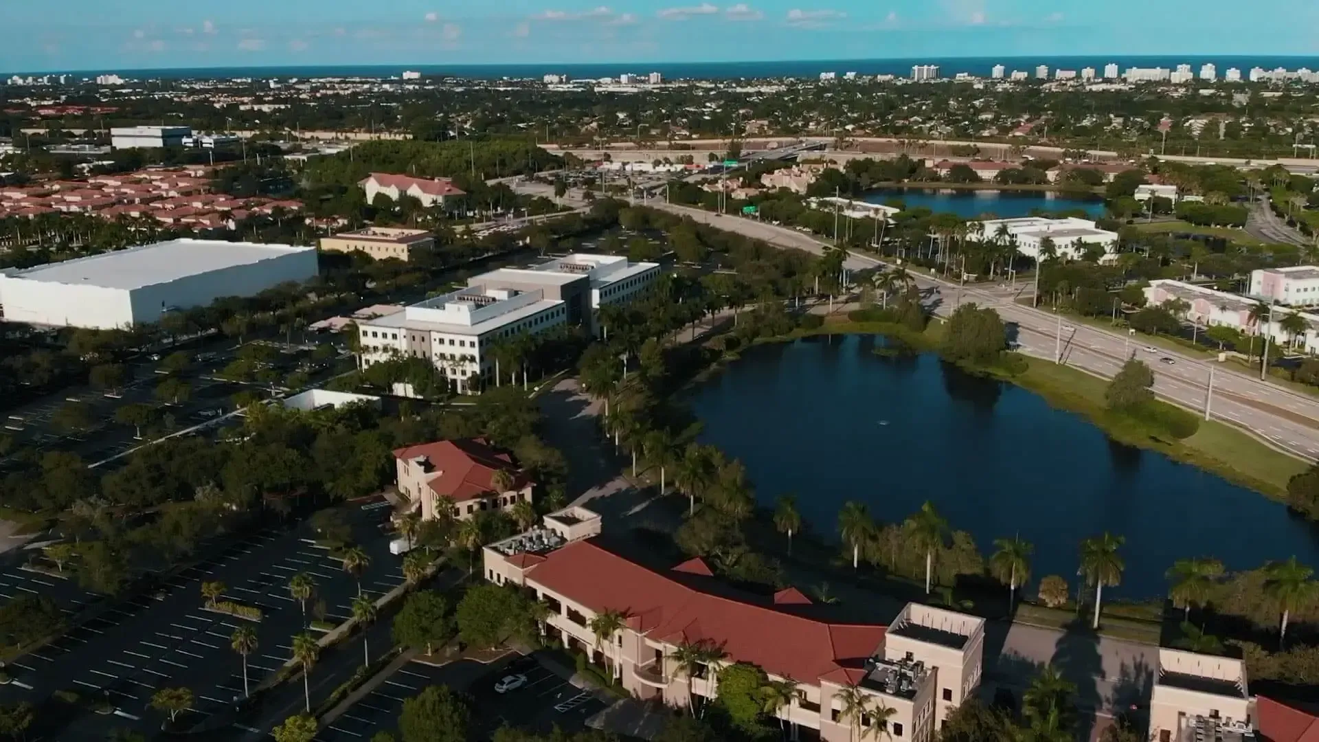 Aerial view of a lakeside neighborhood with red-roofed buildings, palm trees, roads, and a distant city skyline