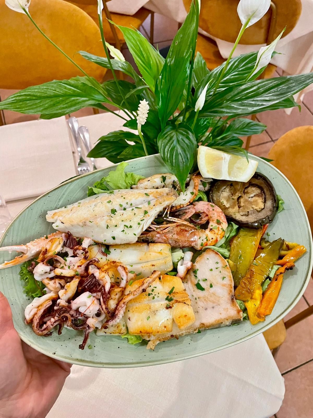 Piatto di frutti di mare misti con spicchi di limone, verdure grigliate ed erbe aromatiche su un piatto bianco.