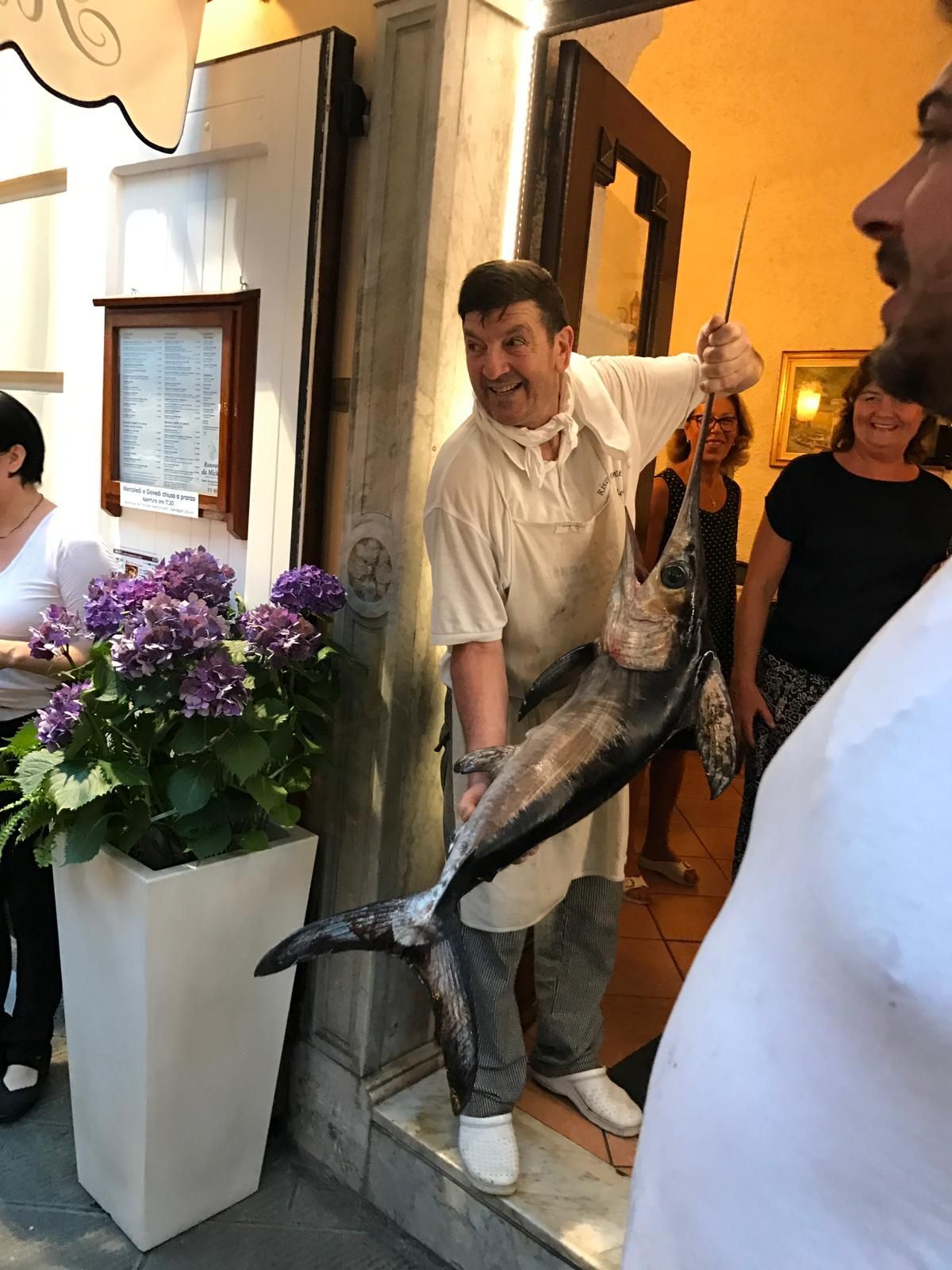 Un uomo tiene in mano un grosso pesce in un ambiente chiuso, accanto a una porta, con fiori e altre persone nelle vicinanze.