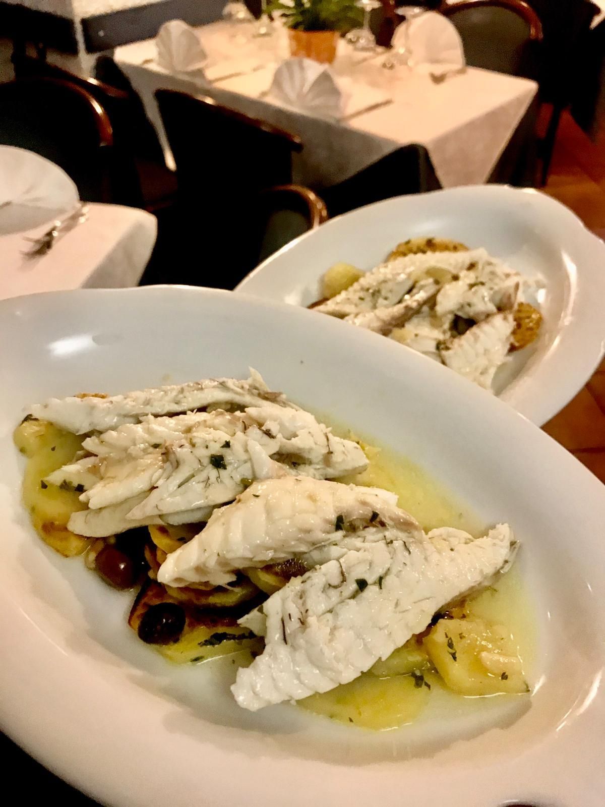 Un piatto di pesce bianco con salsa su un tavolo di ristorante, con un secondo piatto sullo sfondo.