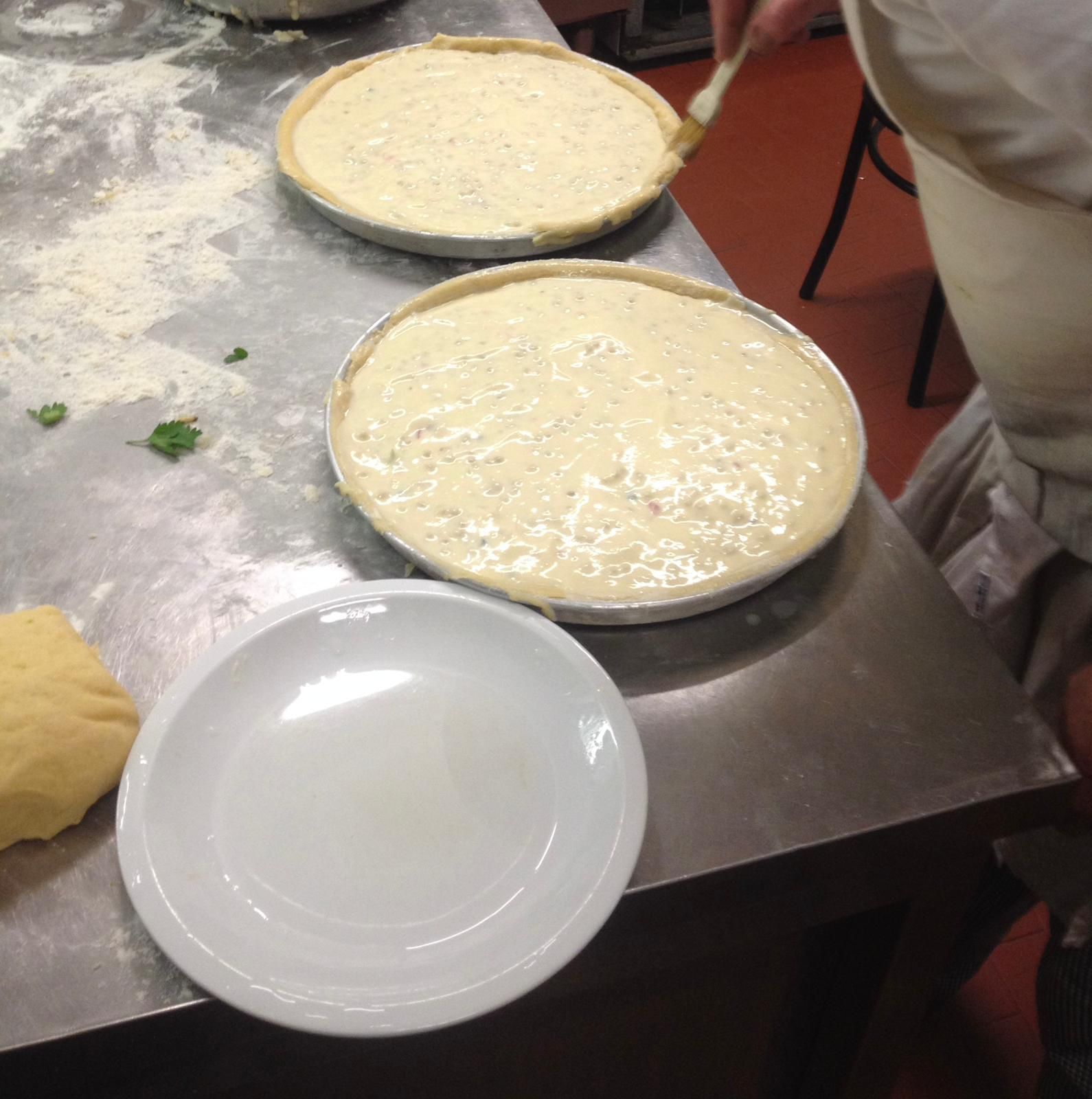 Due teglie per pizza con impasto chiaro su un piano di lavoro infarinato in metallo, accanto a un piatto bianco.