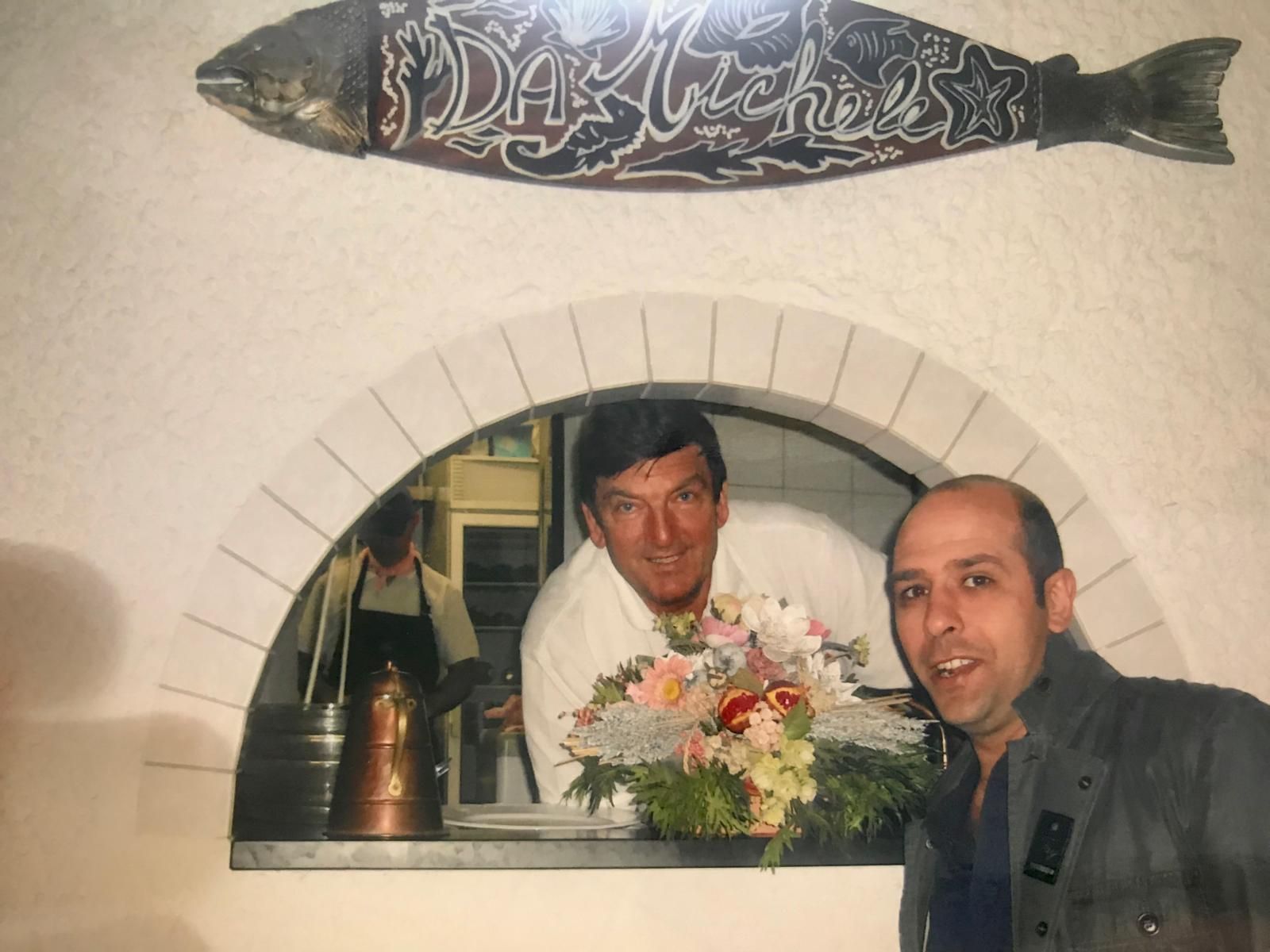 Due uomini sorridenti affacciati a una finestra ad arco in mattoni, con in mano un mazzo di fiori, sormontati da un'insegna decorativa a forma di pesce.