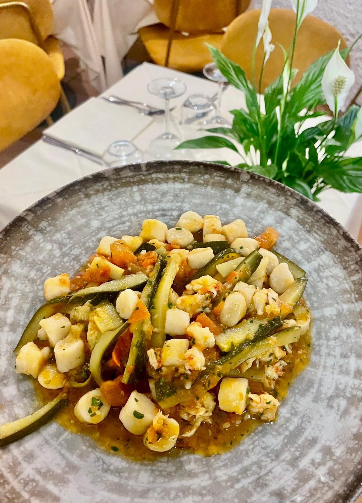 Insalata di verdure grigliate e gnocchi su un piatto grigio, su un tavolo con tovaglie bianche e piante verdi.