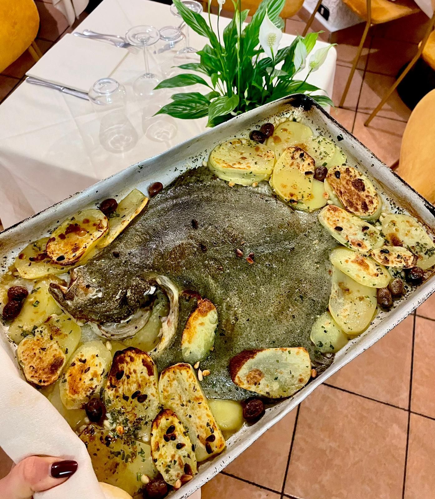 Un vassoio di spicchi di patate al forno con erbe aromatiche e salsa, appoggiato su un tavolo piastrellato con bicchieri e una pianta in vaso.