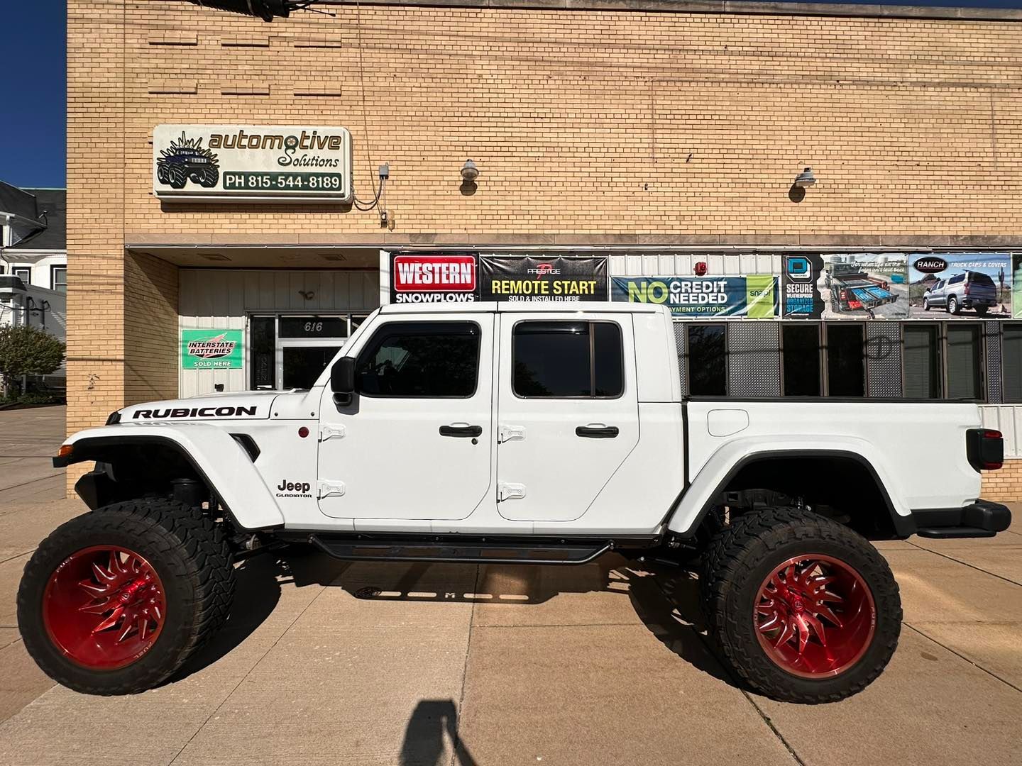 White lifted Jeep Gladiator with red rims parked in front of a tan storefront.