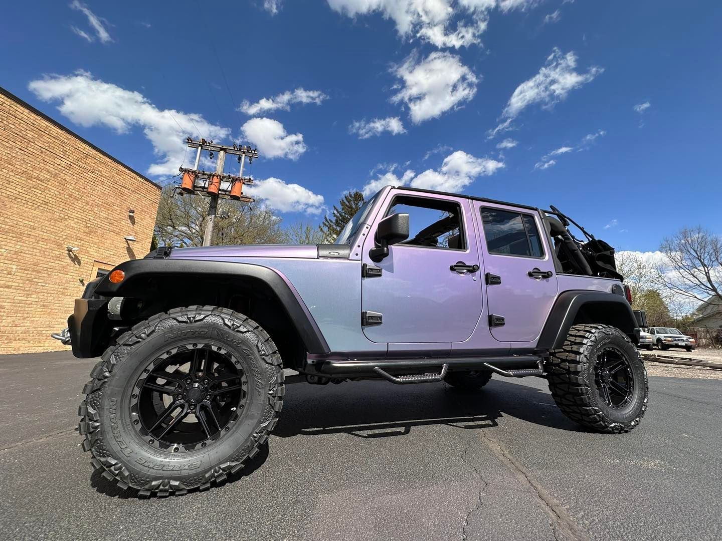 Purple Jeep Wrangler with oversized off-road tires parked on asphalt near a brick building under a blue sky