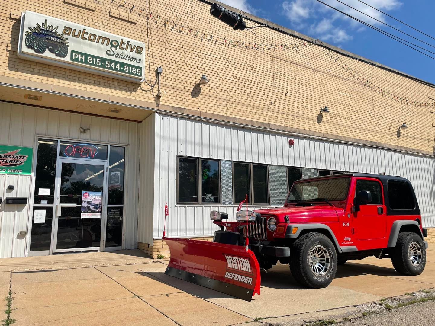 Red Jeep with snowplow parked outside an automotive shop storefront