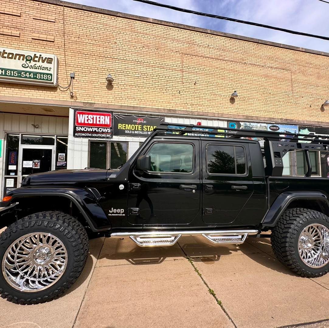 Black Jeep Wrangler parked outside a brick storefront, side view on a sunny sidewalk.