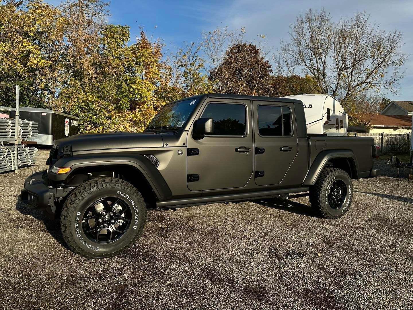 Beige Jeep Gladiator parked on gravel with trees and houses in the background.