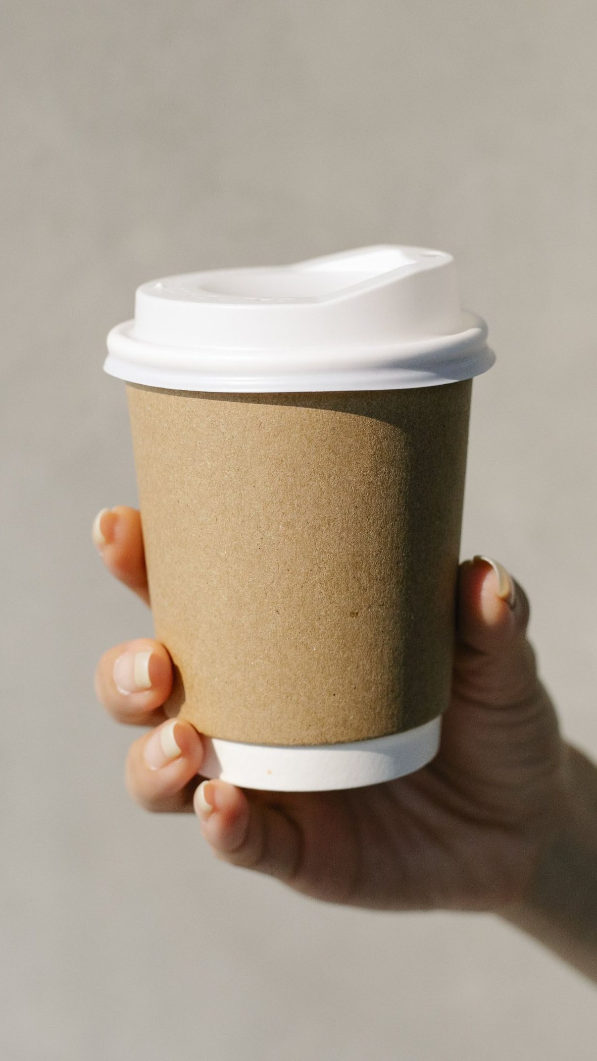 Hand holding a brown paper coffee cup with a white lid against a light gray background.