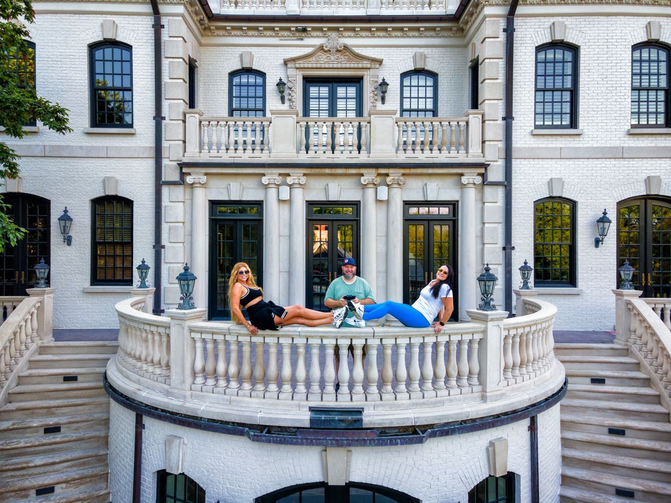 Three people relax on a curved balcony of a large white mansion. Two women and a man pose casually, enjoying the view.
