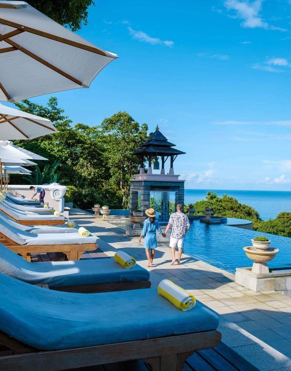 Couple walking by an infinity pool at a resort overlooking the ocean on a sunny day. Lounge chairs and umbrellas are visible.