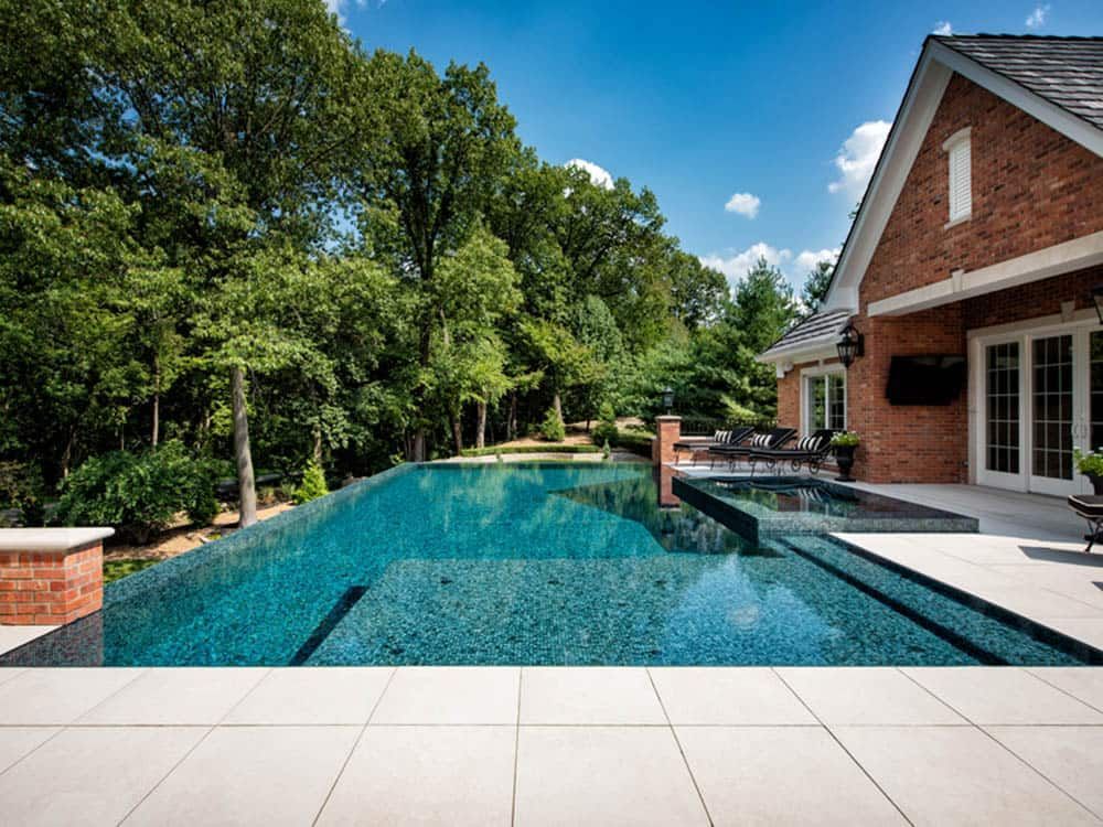 A luxurious backyard pool with a mosaic tile finish sits next to a brick house on a sunny day. Lush trees line the background.