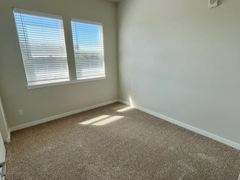 An empty bedroom with a carpeted floor and two windows.