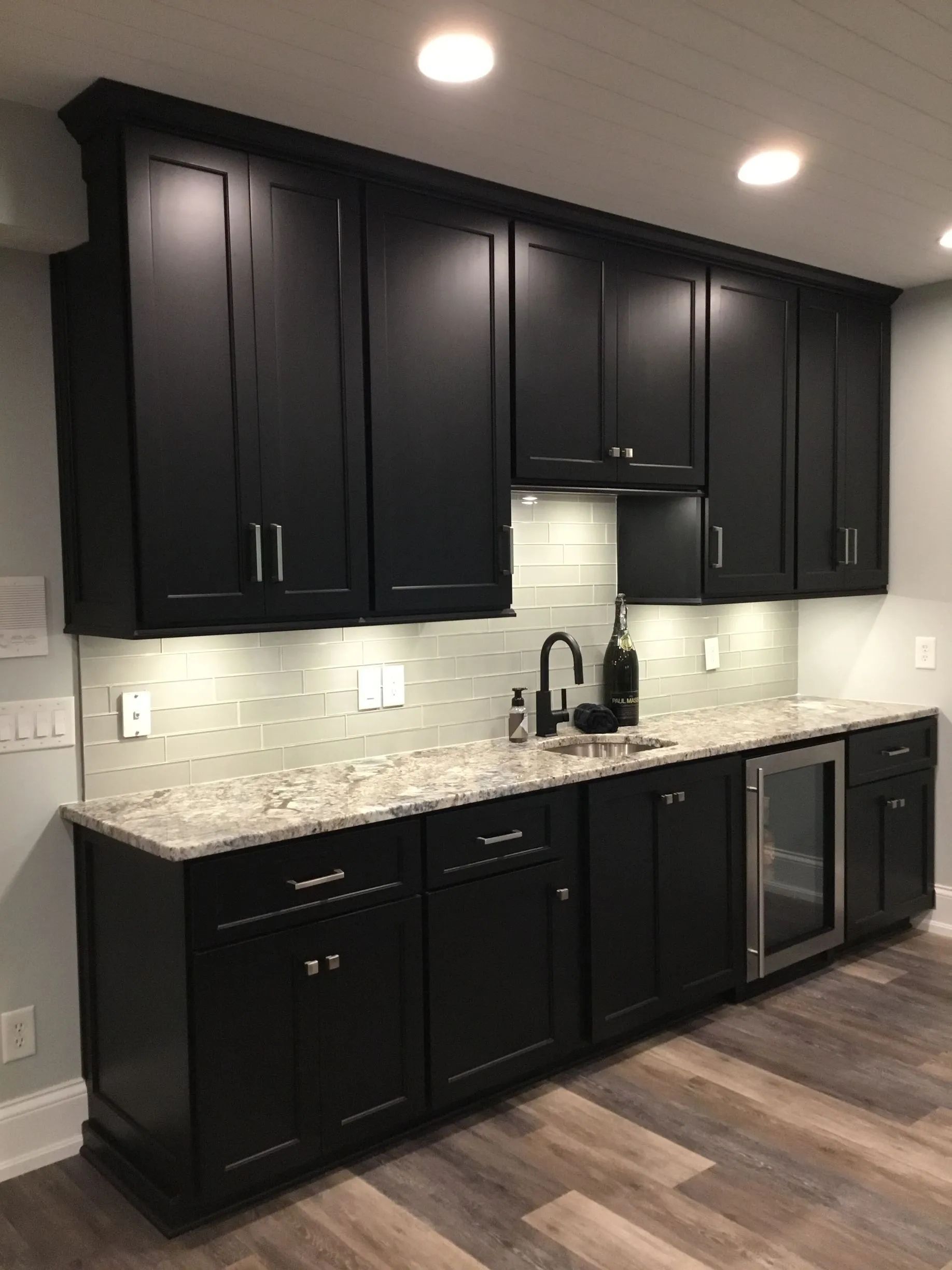 A modern wet bar featuring matte black cabinetry, a light stone countertop, subway tile backsplash, and a wine cooler.