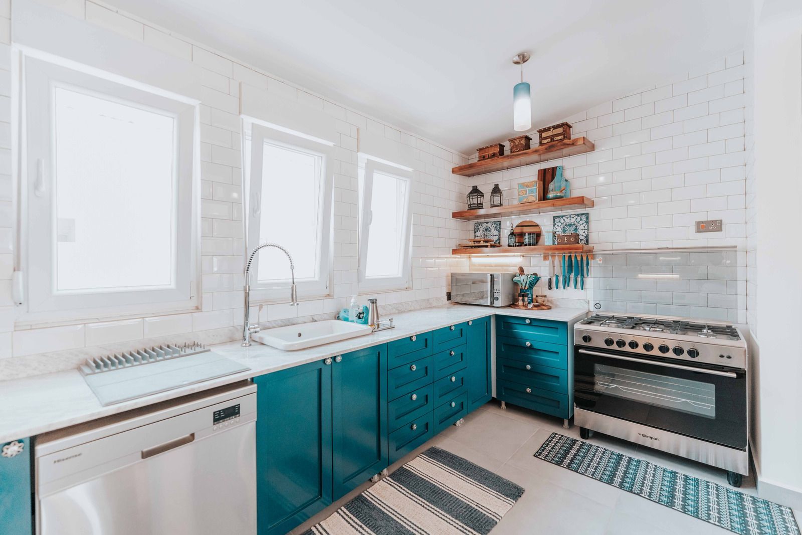 Teal cabinets and stainless steel appliances in a bright kitchen with exposed brick wall and wooden shelves.