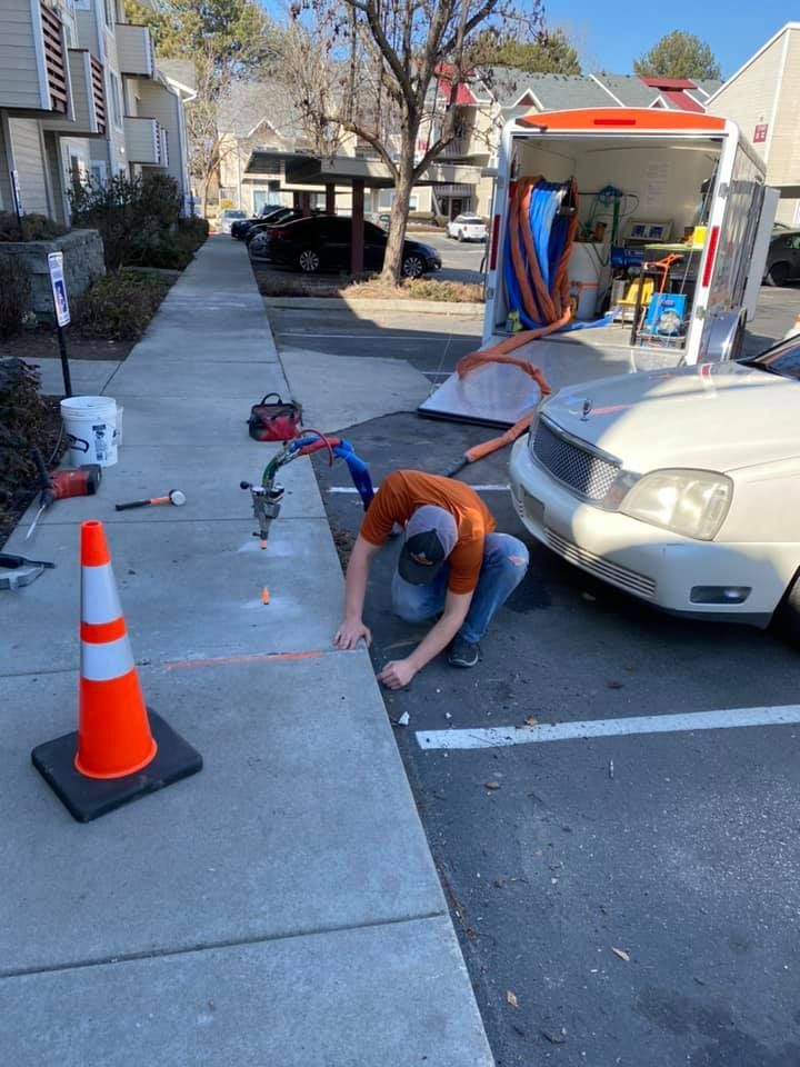 A person crouches, working on a sidewalk near a parked vehicle and utility truck, with orange safety cones.