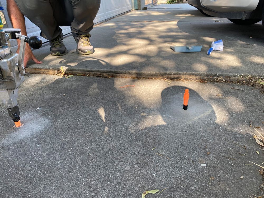 Person kneeling, cutting concrete driveway with a tool. An orange marker and debris are visible.