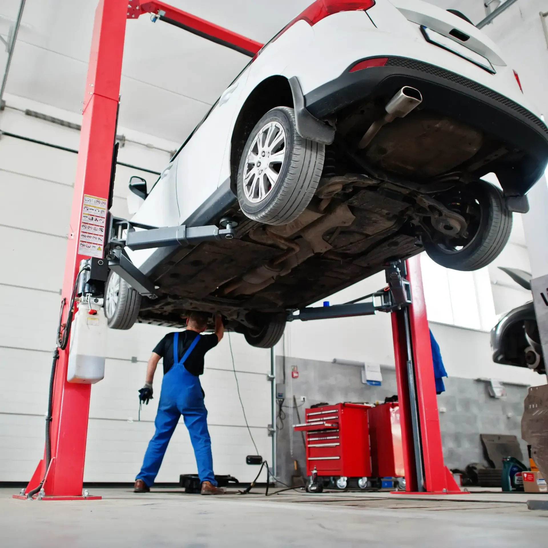 Mechanic in blue overalls inspecting a white car lifted on a red hydraulic hoist in an auto shop