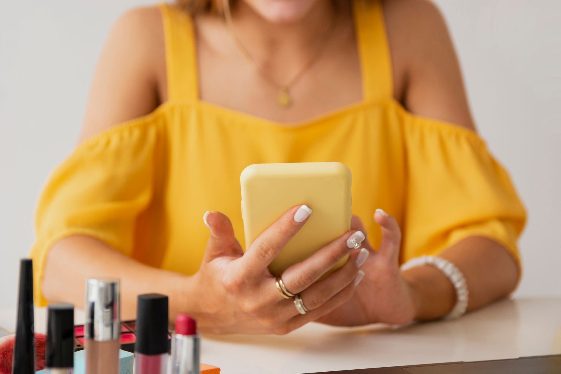 Mulher com blusa amarela segurando celular ao lado de itens de maquiagem em uma mesa.