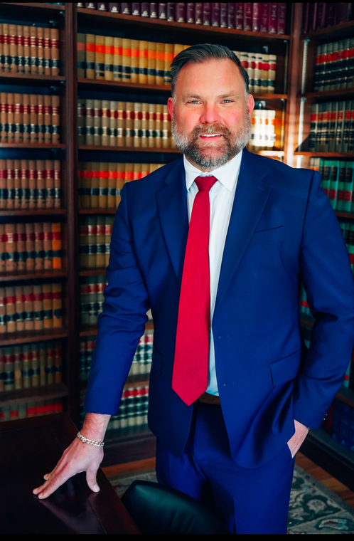 Man in a blue suit and red tie standing in a law library, hand on a chair.