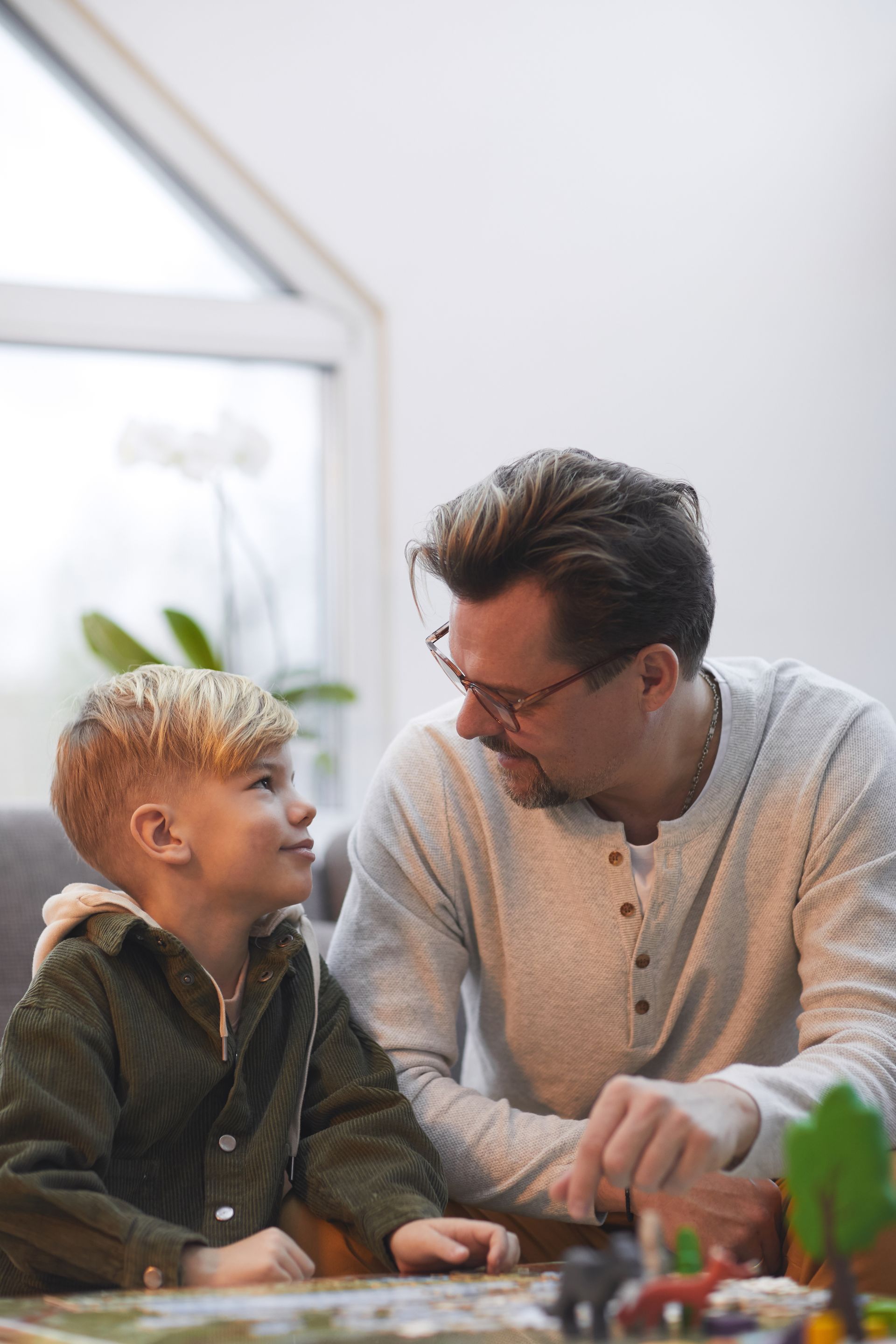 Man and child smiling at each other across a table in a bright room, with a plant in the foreground