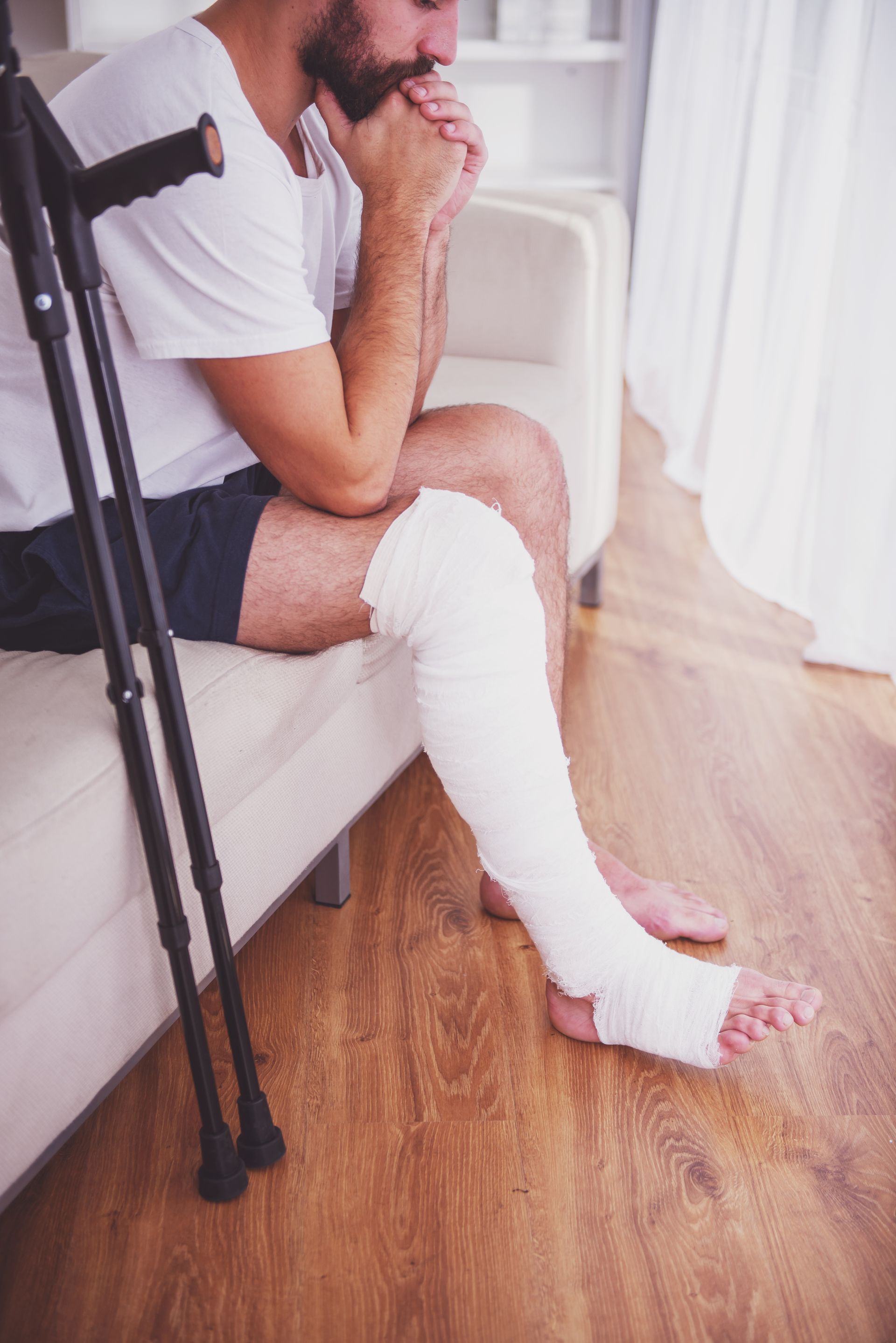 Man seated indoors with a white leg cast and crutches beside him, resting with hands clasped.