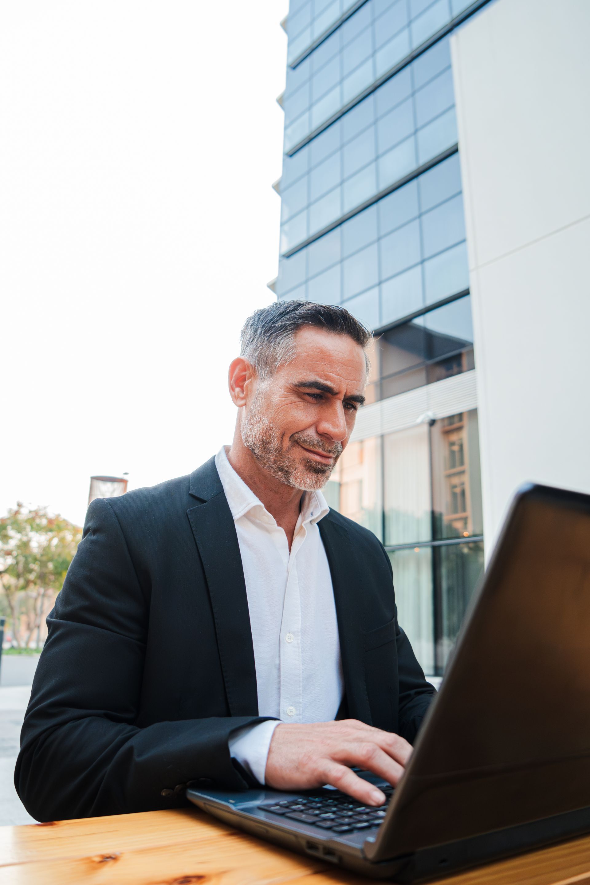 Man in a black blazer using a laptop outdoors beside a modern glass building