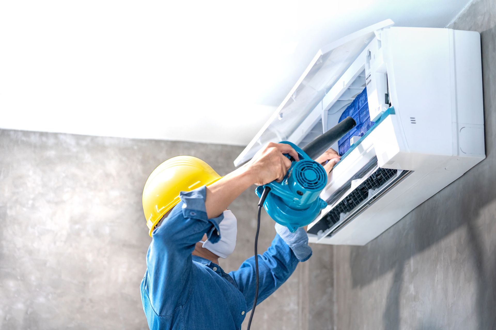 Technician in a yellow hard hat repairing a wall-mounted air conditioner with a drill.