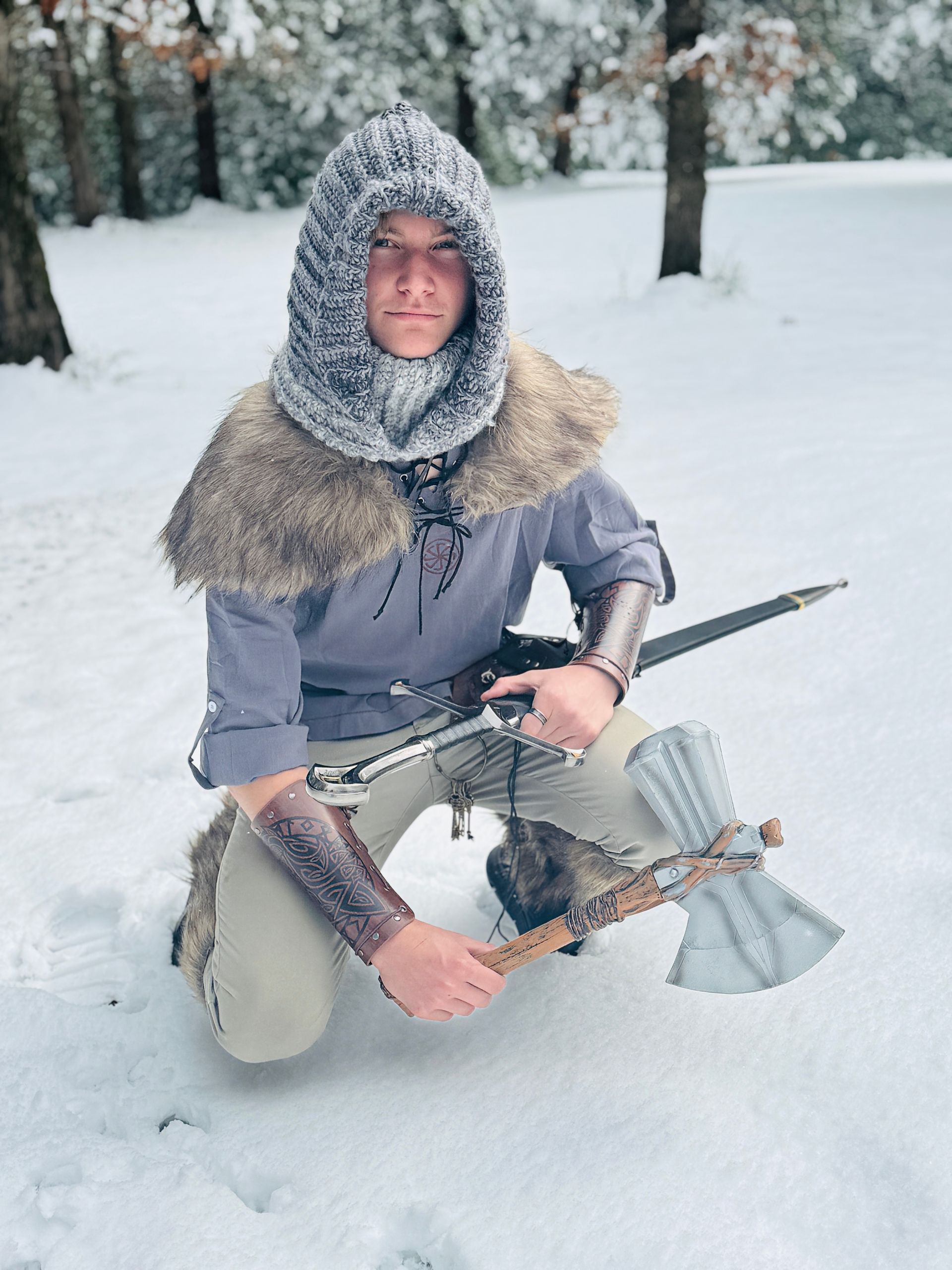 Person in medieval garb kneels in snow, holding an axe and a bow, wearing a fur-trimmed hood.
