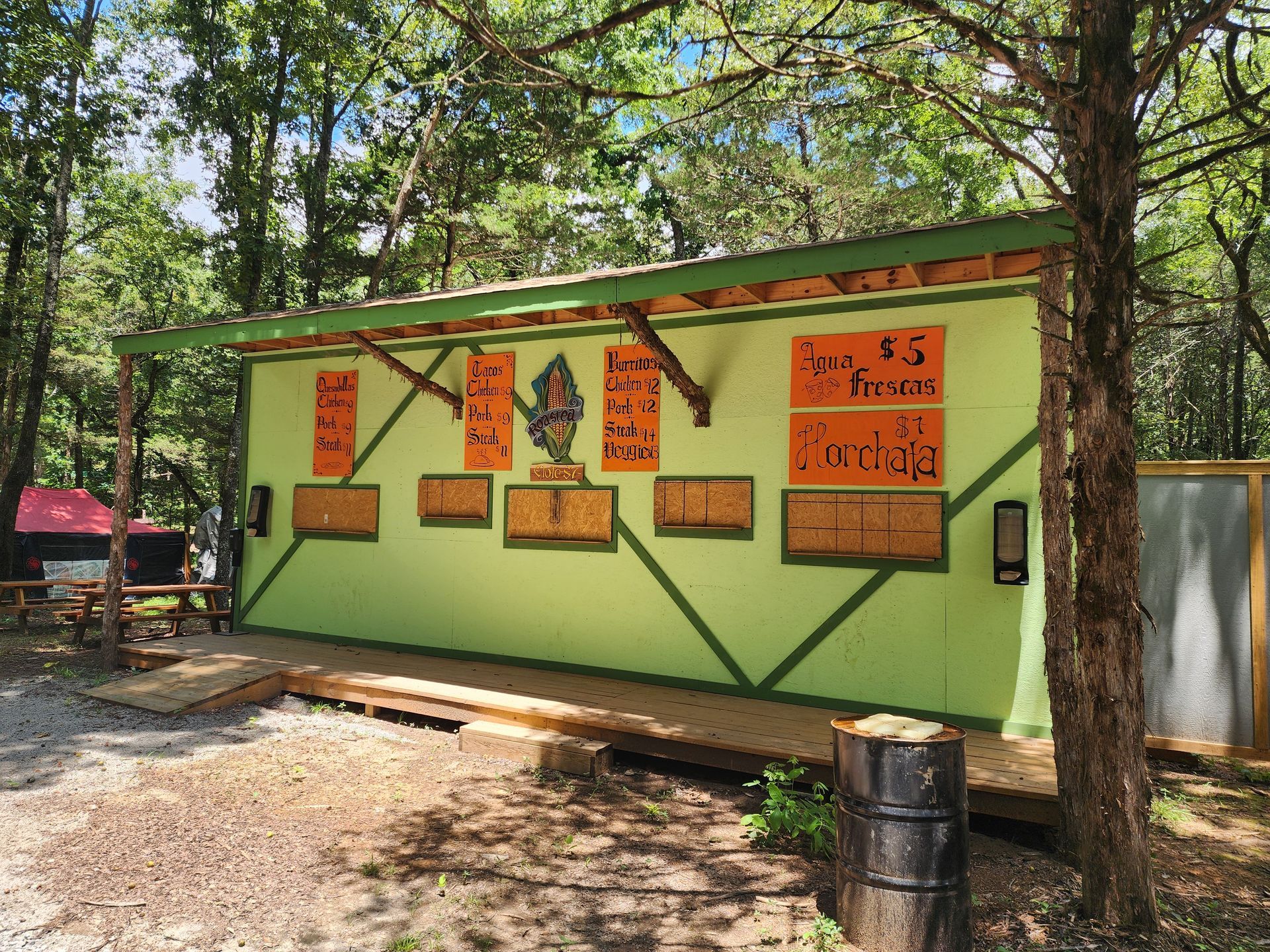 Small green food stand with orange menu signs in a wooded outdoor area