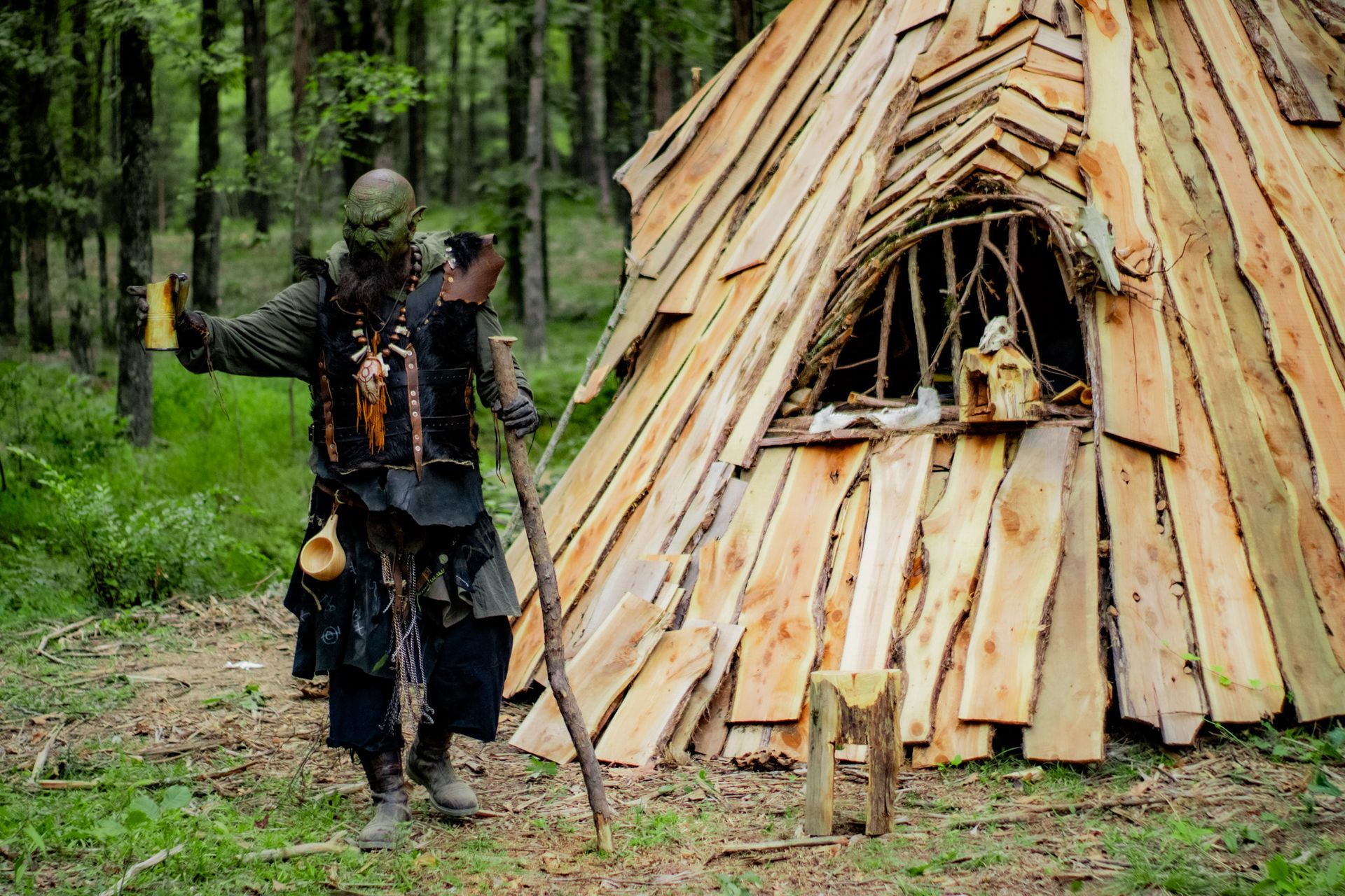 A man in a orc costume is standing in front of a wooden hut in the woods.